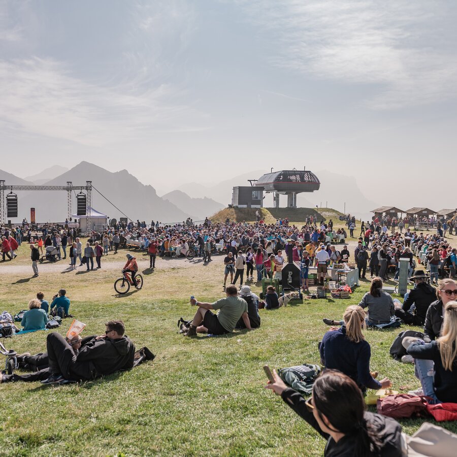 Besucher sitzen entspannt im Gras vor der Bühne bei sonnigem Wetter und genießen das Fest. | © Gianvito Coco