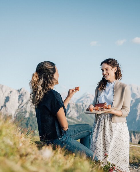 Vor dem Bergpanorama sieht man eine stehende Frau mit einem Brett Speck in den Händen und sie lächelt eine sitzende Frau an, welche sich an dem Speck bedient. | © IDM Südtirol