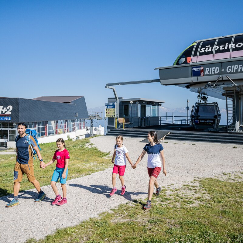 Eine Familie mit 2 Kindern steigt aus der Bergbahn aus und läuft bei Sonnenschein den Weg entlang. | © Wisthaler
