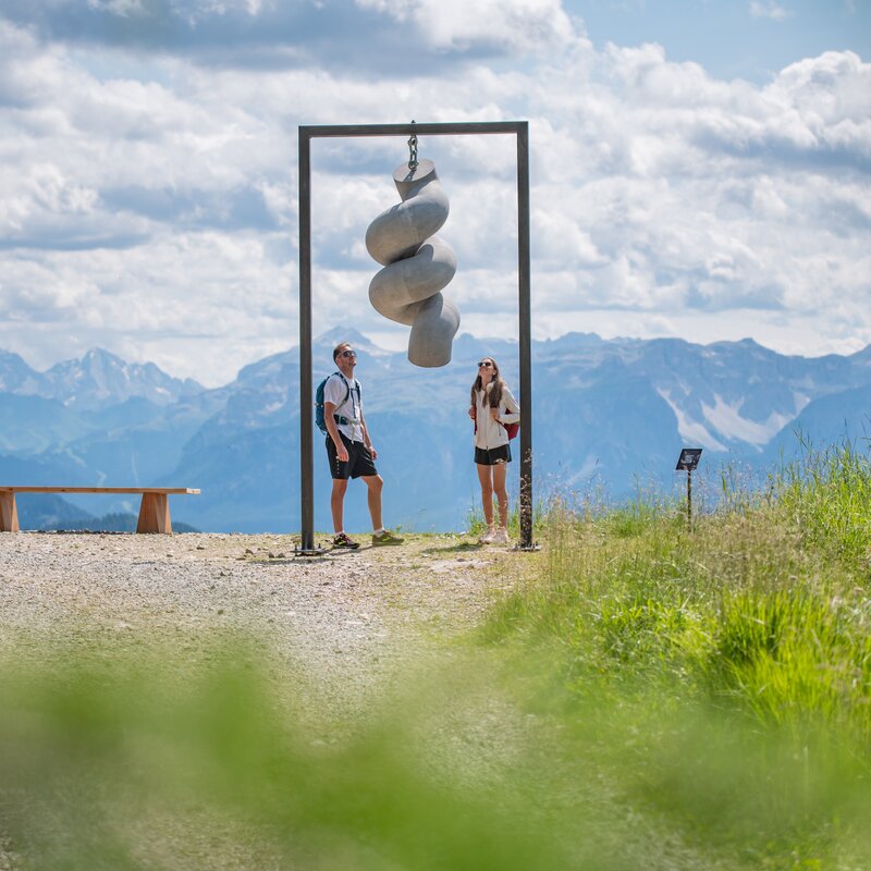 Zwei Personen stehen unter einem hängenden Kunstwerk auf einem Berg, im Hintergrund das Bergpanorama und der leicht bewölkte Himmel. | © Gianvito Coco